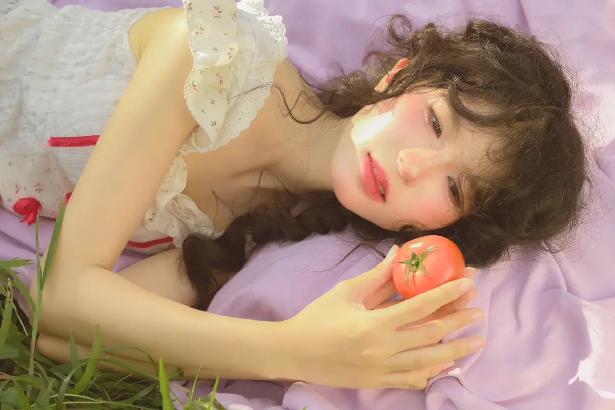 白いドレスを着た女性が紫陽花の生地の上に寝そべっている。熟れたトマトを手に持っている。