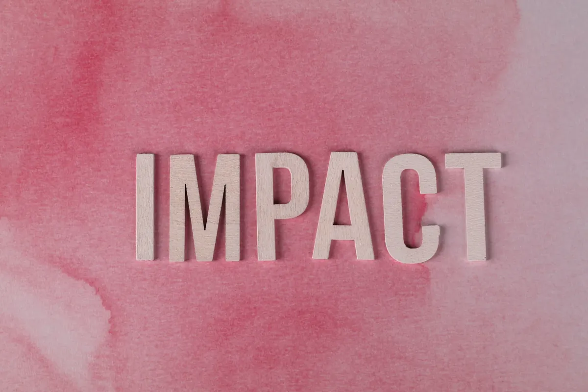 木製の文字「IMPACT」が、質感のあるピンクの水彩画の背景に描かれている。影響と変化を象徴している。